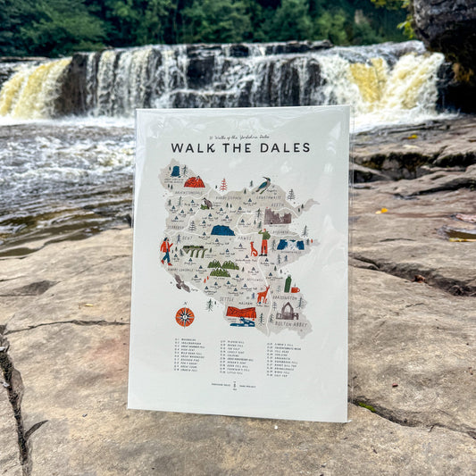 A white illustrated map poster titled "WALK THE DALES" stands upright on a rock in front of a waterfall and forest. The poster features a stylized map with small colorful icons and a list of locations below.