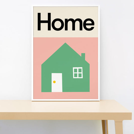 Framed poster with a green house illustration and 'Home' text on a pink background, placed on a wooden surface.