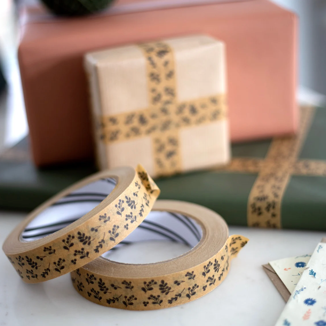 Botanical patterned washi tape rolls on a white marble surface, with wrapped gift boxes in the soft-focus background.