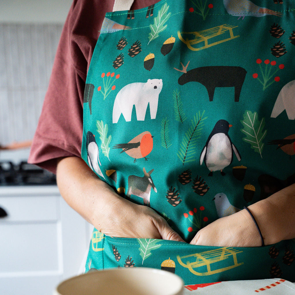 A person wearing a green apron with a winter animal and sledge print, standing in a kitchen and placing their hands in the pocket of the apron. A striped mug sits on a tray in the foreground.