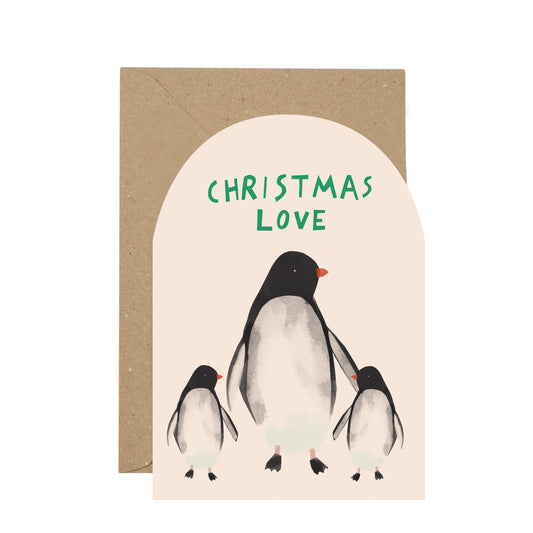 A Christmas greeting card with an arched cream front featuring three penguin illustrations and the words 'CHRISTMAS LOVE' in green, shown with a brown kraft envelope behind it on a white background.