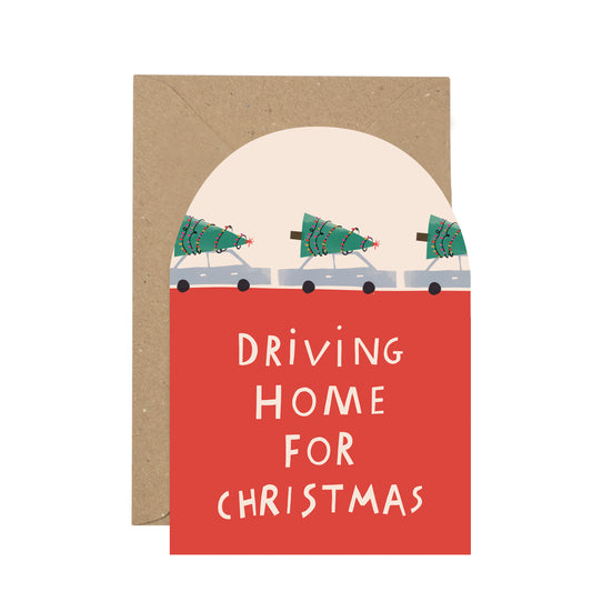 A holiday greeting card with a kraft envelope behind it. The card has a red lower section with white text reading “DRIVING HOME FOR CHRISTMAS,” above an illustration of three Christmas trees in cars under a cream semicircle.