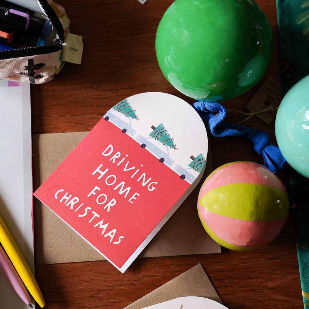 A festive Christmas greeting card with the text 'DRIVING HOME FOR CHRISTMAS' in white on a coral-red background, styled on a wooden desk alongside green and pastel decorative balls and stationery.