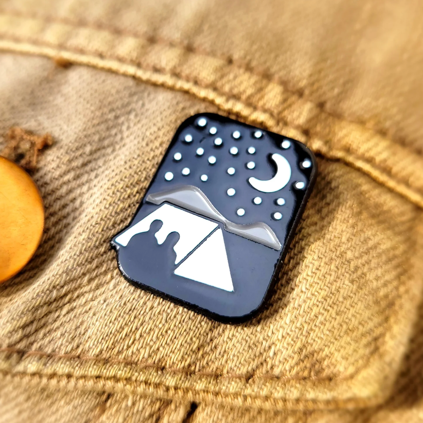 Enamel pin with a design of two people under a tent under a starry night sky on a denim background.