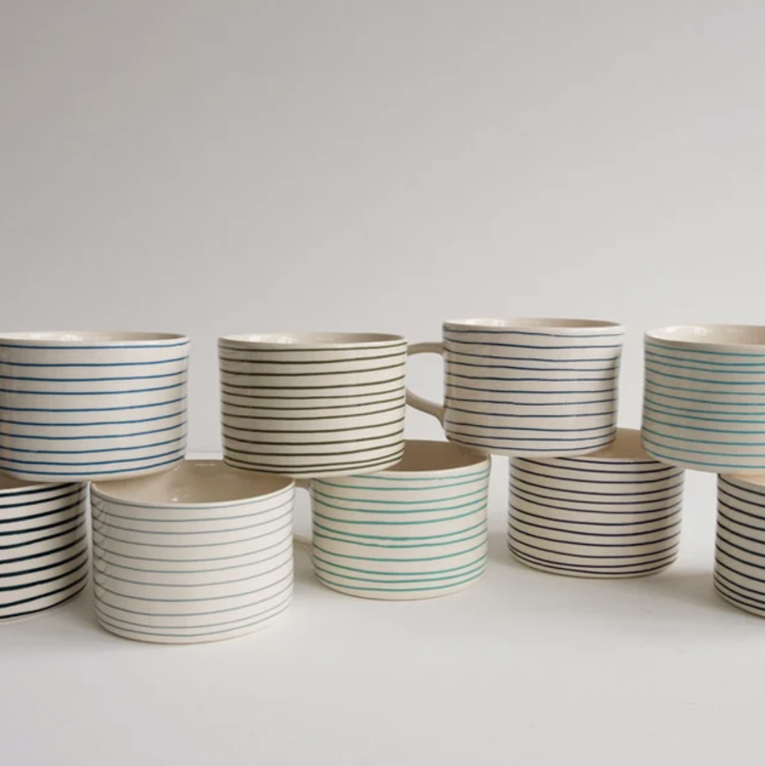 A group of ceramic mugs with thin horizontal stripes in various colors, including blue, green, and black on an off-white base, arranged on a light gray surface against a neutral light gray background.