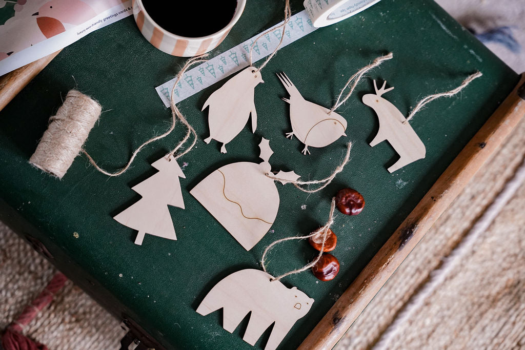 A set of natural wood Christmas ornaments shaped like penguins, a Christmas tree, a reindeer, a polar bear, and a snowman, arranged on a dark green textured surface with twine, thread, and small red beads.