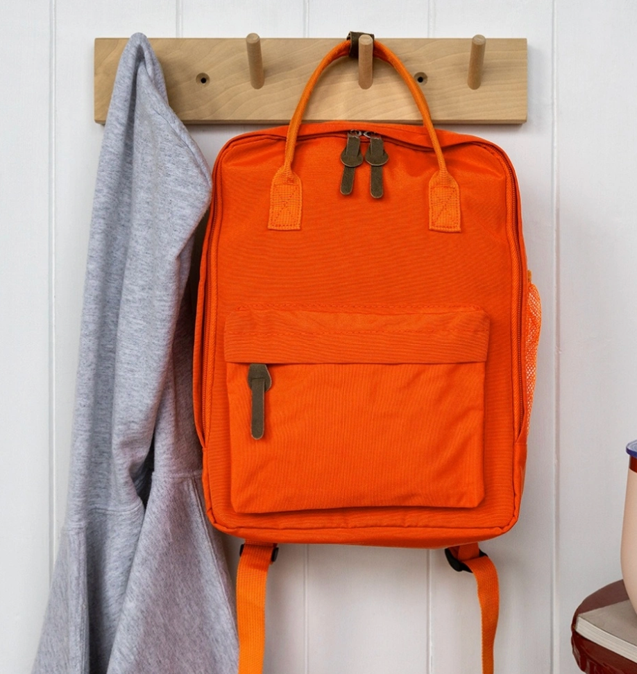 An orange backpack with dual top handles and a front zip pocket hangs from a wooden peg rack on a white paneled wall, next to a gray hoodie.