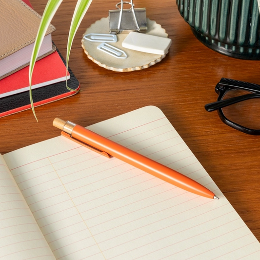 An orange pen with a wooden accent lies on an open lined notebook on a wooden desk, surrounded by office items including a plant, binder clips, a small eraser, and eyeglasses.