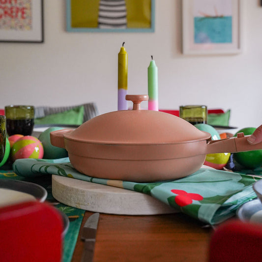 an image of a pink our place ceramic pan placed on a berries christmas tea towel, in a christmas table setting with two colourful candles in the background