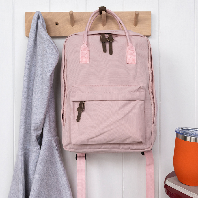 Pink backpack hanging on a wooden hook with a gray sweater and orange tumbler in the background.