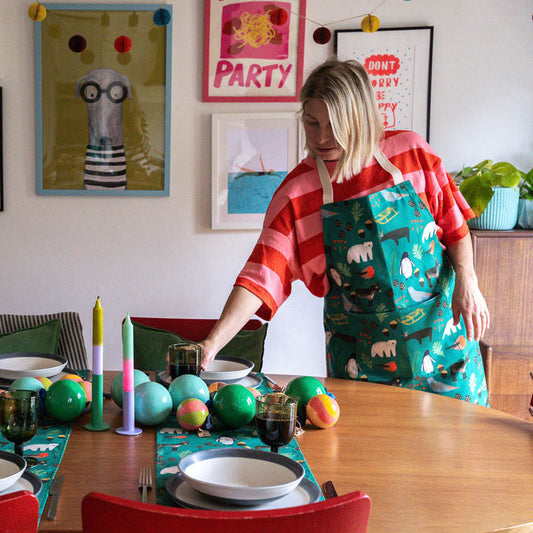 A woman wearing a red and white striped shirt and a green Christmas themed apron with a polar bear pattern is setting a table with colourful christmas baubles, placemats, and candles in a decorated room with framed art.