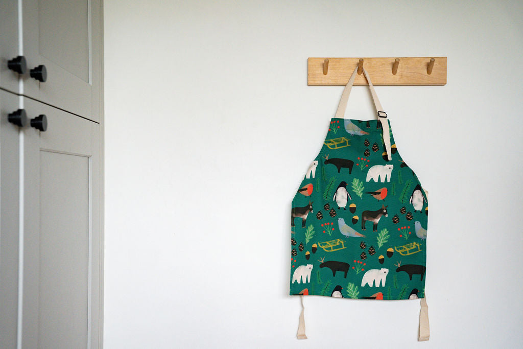 A green childrens kitchen apron with a whimsical animal print hangs from a wooden peg on a white wall, next to a grey cabinet.
