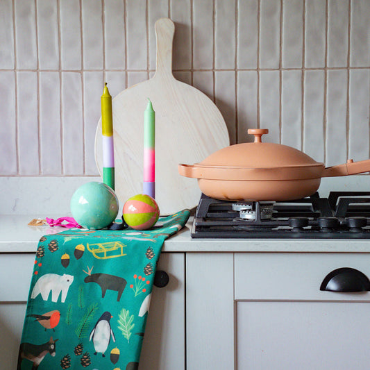 A colourful kitchen scene featuring a green kitchen towel with a whimsical animal print hanging from a white drawer, next to a terracotta-coloured pan on a gas stovetop. A wooden paddle board leans against a beige tiled backsplash in the background.