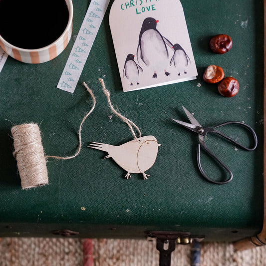A styled flat-lay of Christmas craft supplies on a dark green surface, including a penguin ornament card reading 'CHRISTMAS LOVE', twine, scissors, a spool of thread, and a coffee mug.
