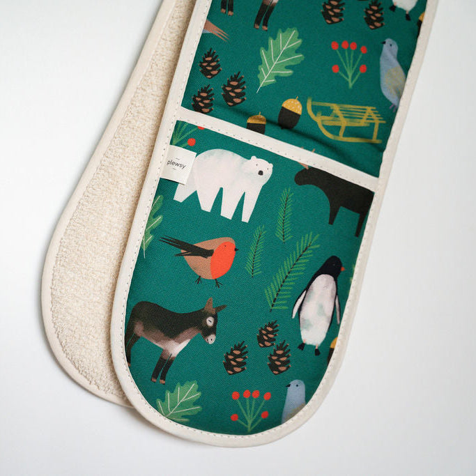 A close-up of a fabric oven mitt or pot holder with a winter animal print on a teal background, showing a cream-colored textured reverse side, set against a plain white background.