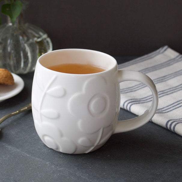 White textured mug with tea on a dark surface with cookies and a vase in the background