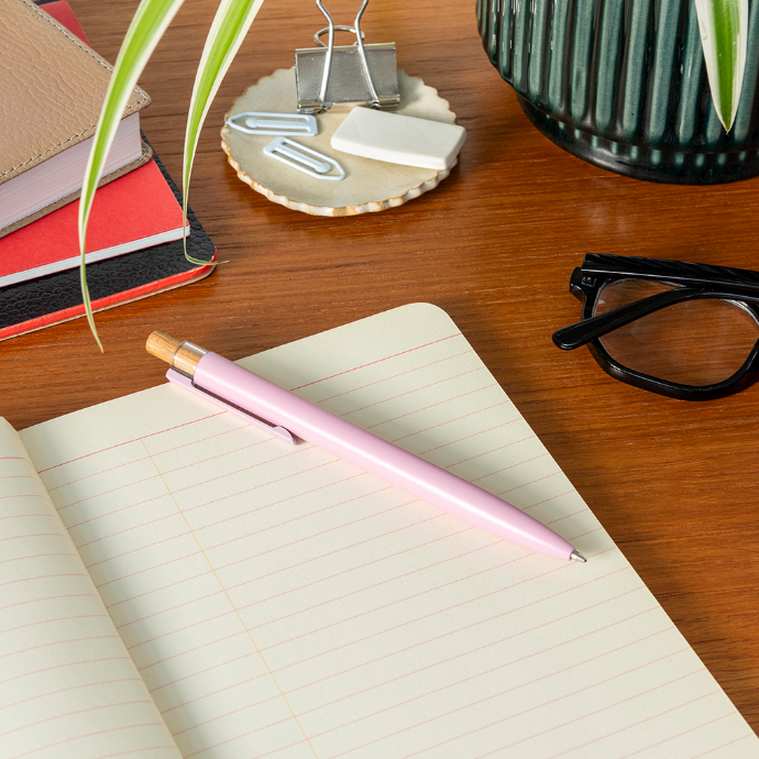 Pink pen on an open notebook with stationery items on a wooden desk
