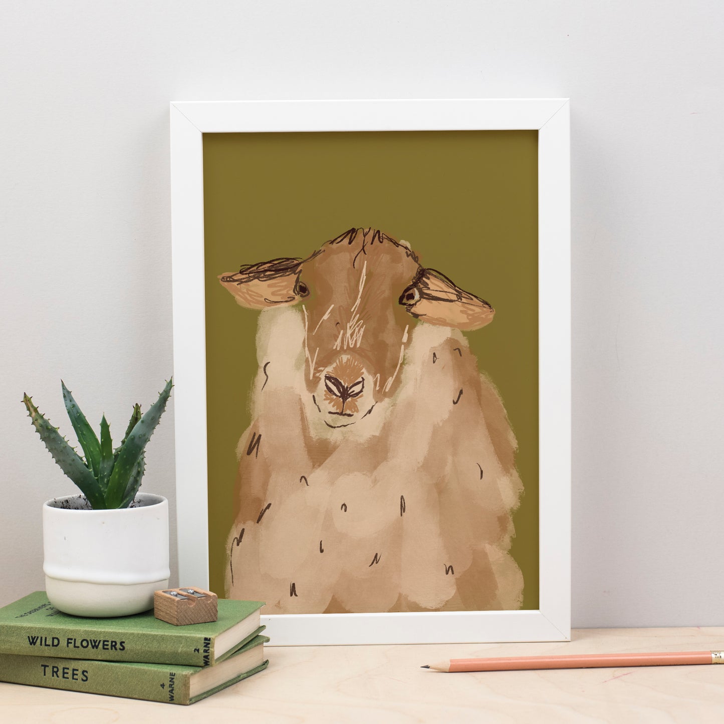 A sheep print featuring an illustration of a sheep on a green and brown background, framed in a white enclosure, displayed on a table with books and a pencil.