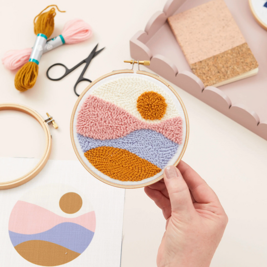 Hand holding a completed embroidery hoop with a colorful landscape design, surrounded by crafting materials on a light background.