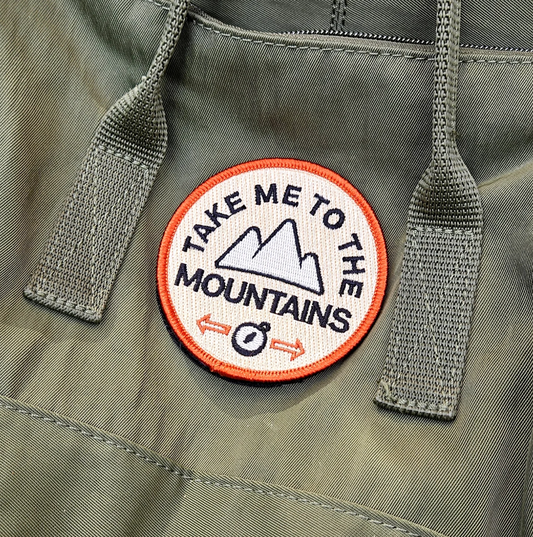 Patch on a jacket with 'Take Me to the Mountains' text and mountain graphic.