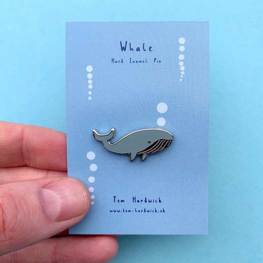Whale enamel pin held by a hand on a blue card with branding details.