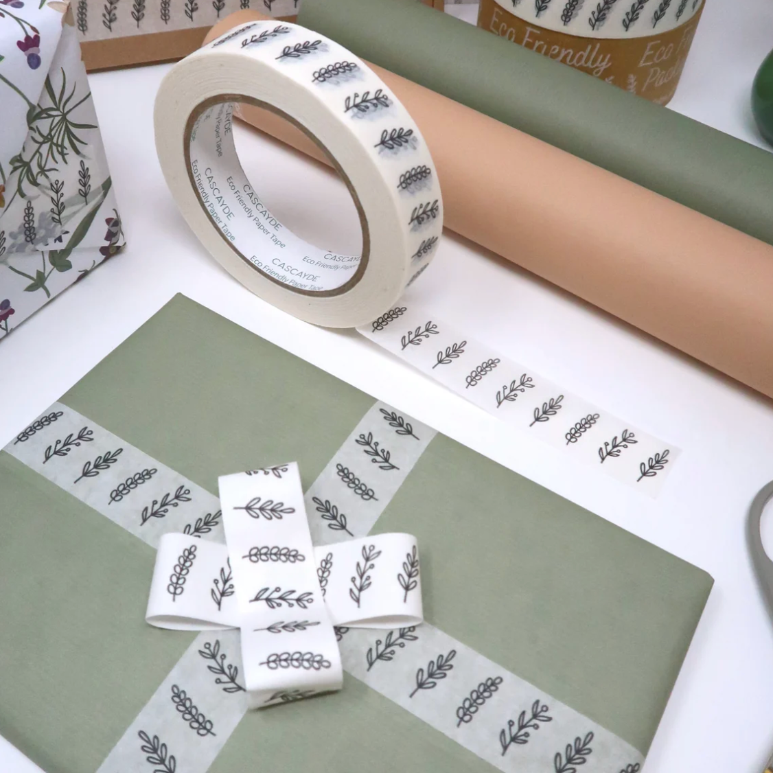 A roll of decorative washi tape with a repeating gray botanical leaf pattern on a white background, shown alongside wrapped gift boxes in muted green and tan tones on a white tabletop.
