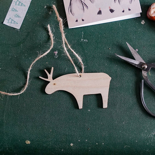 A styled flat-lay featuring a wooden reindeer ornament hanging from twine on a dark green suitcase, surrounded by a mug of black coffee, a measuring tape, a penguin card reading 'Christmas Love', scissors, and dried red fruits.