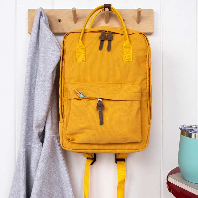Yellow backpack hanging on a wooden hook with a gray jacket and blue tumbler in the background.