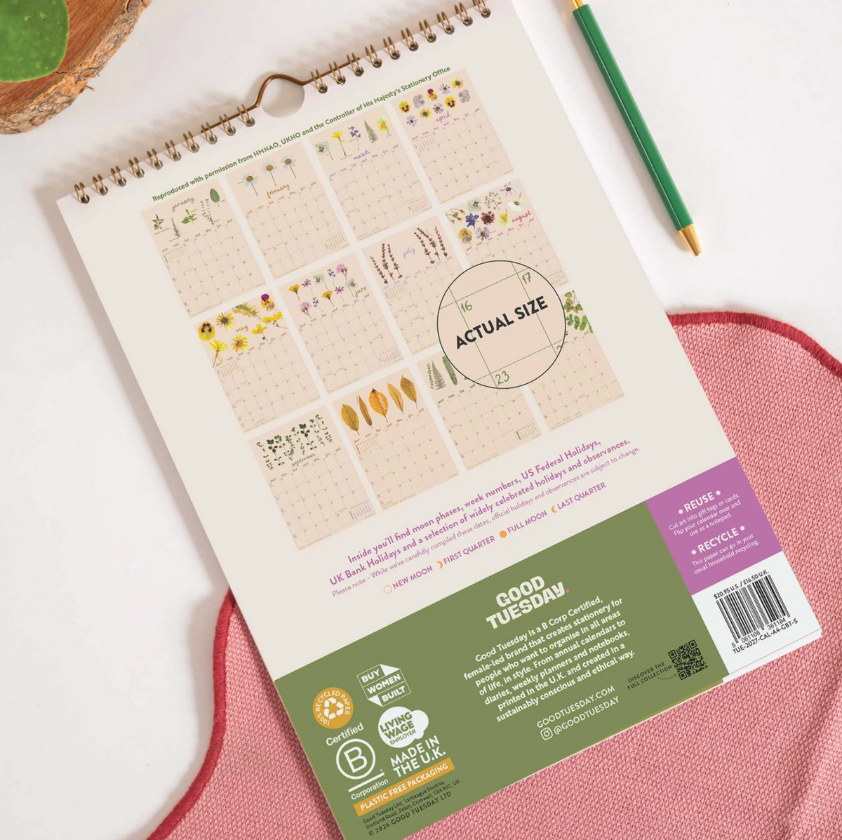 A spiral-bound planner or calendar shown from the back, laid flat on a white surface with a green pencil and a pink textured mat. The page displays a grid of weekly planner templates and a large 'ACTUAL SIZE' circle, with a green and white footer section containing product details and recycling icons.