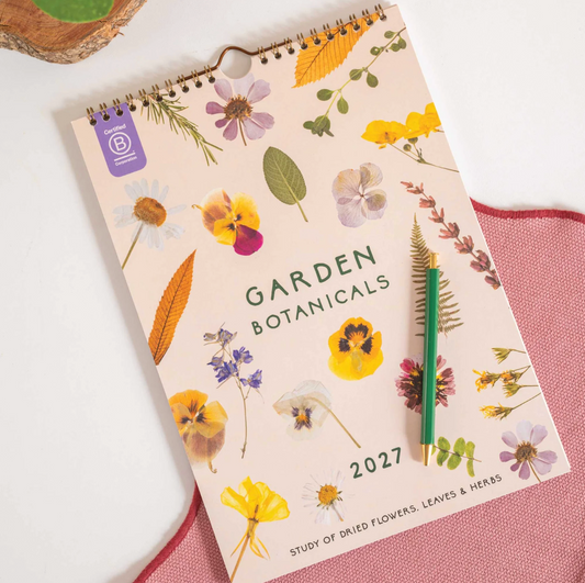 A spiral-bound 2027 Garden Botanicals 2027 calendar lies open on a pink textured surface, showing a floral cover design with pressed flowers, leaves, and herbs. A green pencil rests on the cover.