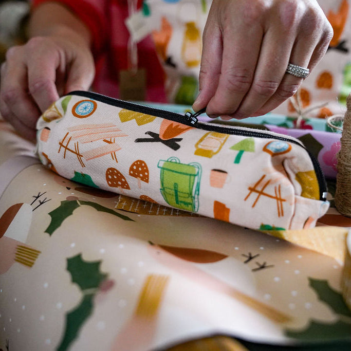 A close-up of a person's hands holding a zippered pencil case with a colourful camping themed pattern, set against a background of patterned wrapping paper and crafting supplies.
