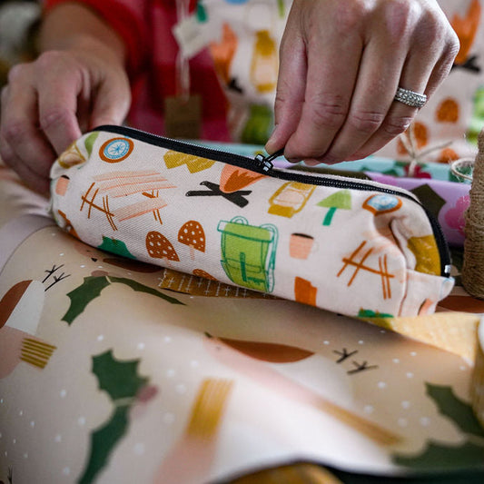A close-up of a person's hands holding a zippered pencil case with a colourful camping themed pattern, set against a background of patterned wrapping paper and crafting supplies.