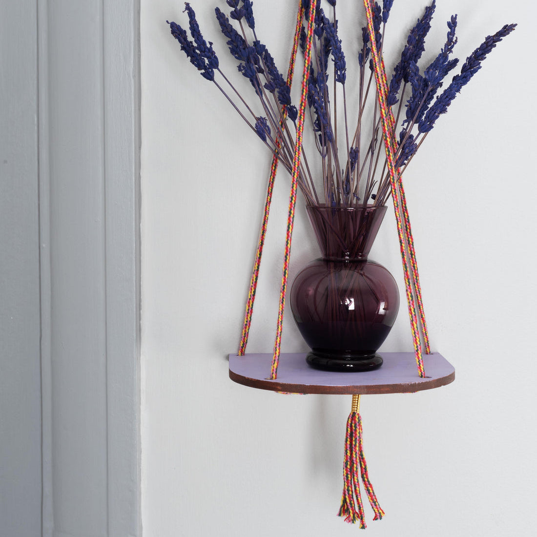 Hanging plant holder with a vase containing lavender against a light gray wall.