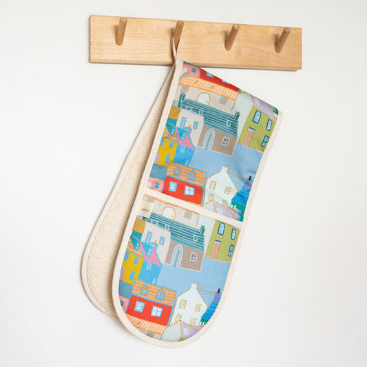 A fabric oven mitt with a colourful coastal house pattern hangs from a light wood wall hook against a plain white wall.