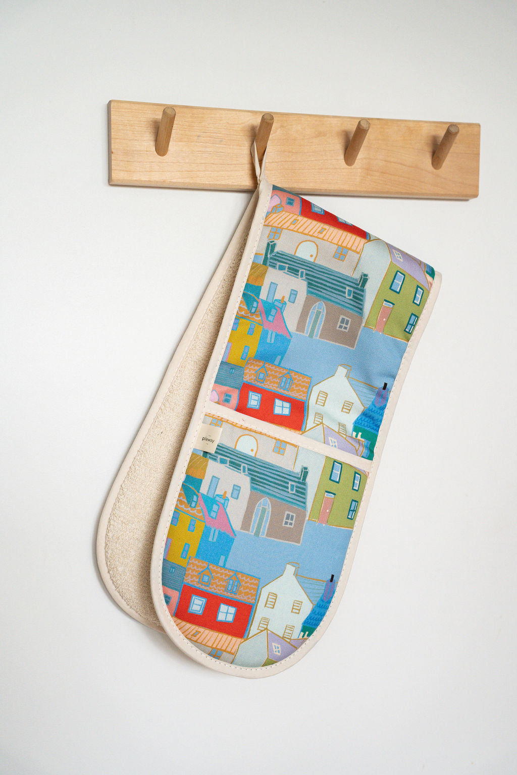A fabric oven mitt with a colourful coastal village illustration pattern, hanging from a light wood peg on a white wall.