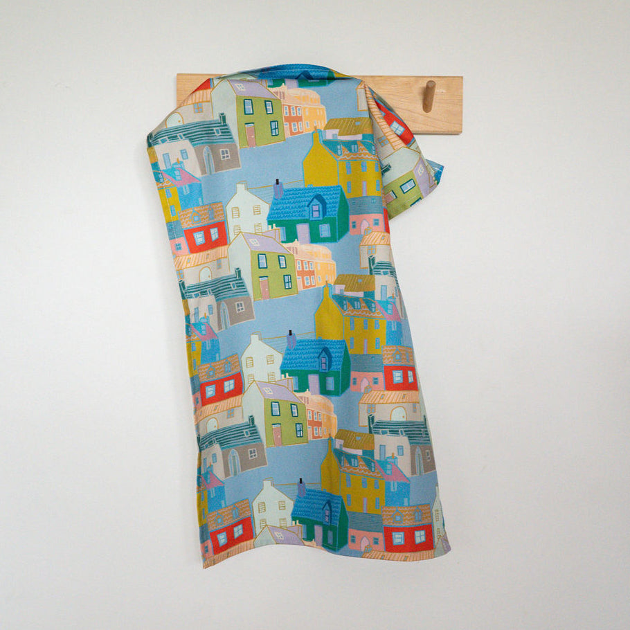 A cotton tea towel with a colourful house pattern, hanging on a wooden wall hook against a white wall.