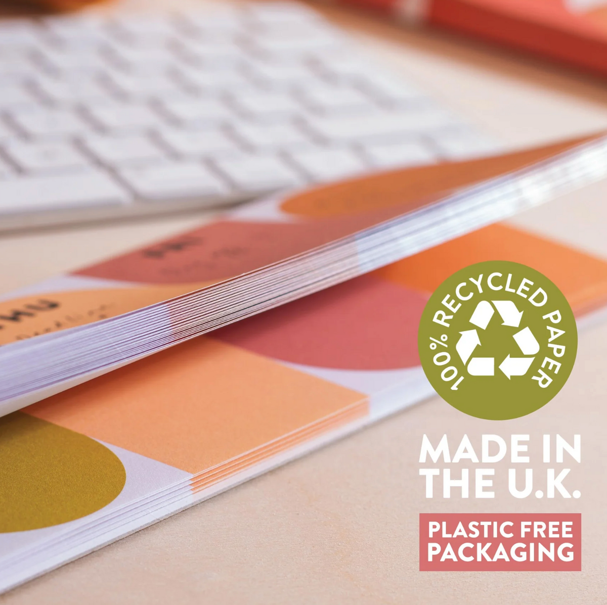 A stack of colorful printed paper packaging or booklets on a desk, with a blurred keyboard in the background. A green circular badge reads '100% Recycled Paper' and text on the right says 'Made in the U.