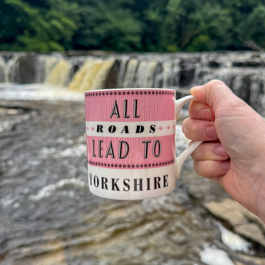 A hand holds a white ceramic mug with pink and black text reading 'ALL ROADS LEAD TO YORKSHIRE' in front of Aysgarth Falls waterfall and forest background.
