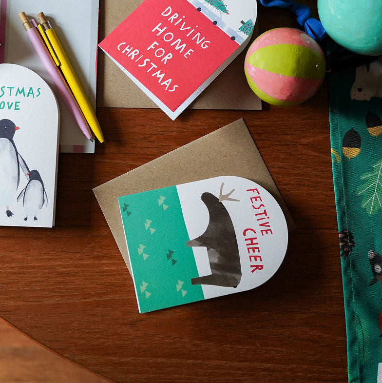 A styled flat-lay of Christmas-themed stationery and craft supplies on a wooden table, including greeting cards, a pencil pouch, washi tape, string, and colorful eggs.