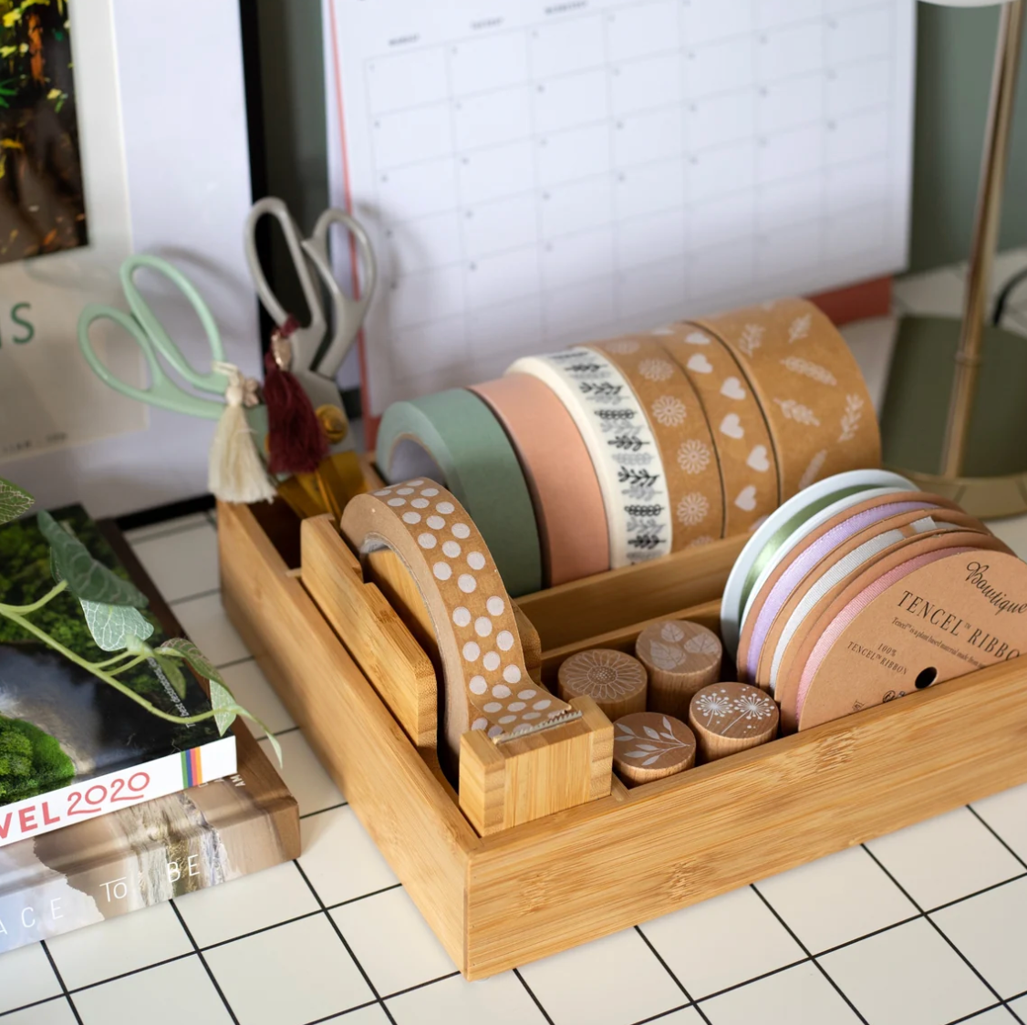 A bamboo desk organizer tray holding rolls of decorative washi tape, pastel paper circles, and small round wooden pieces on a tiled desk, with scissors and a calendar nearby.