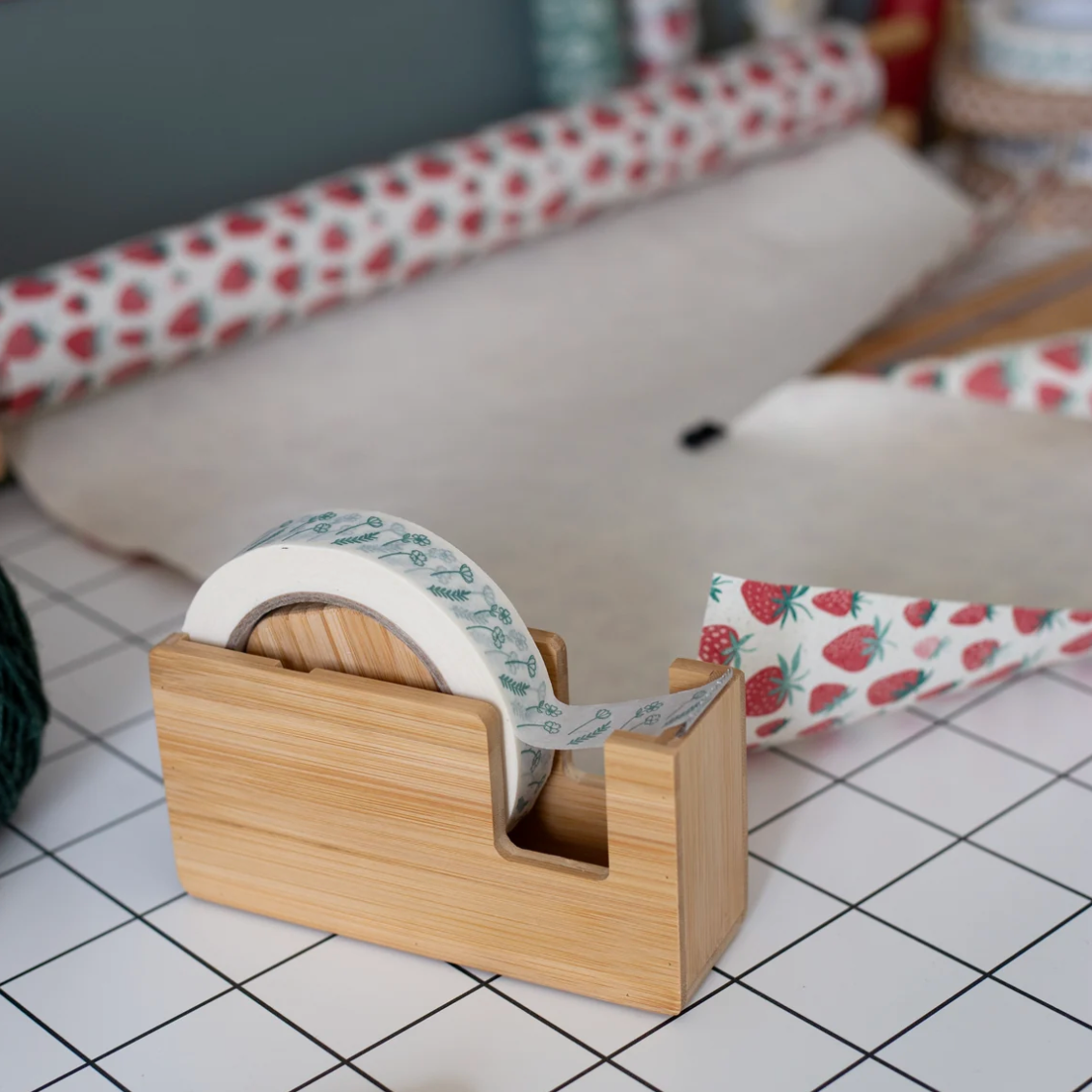 A roll of washi tape with a strawberry and blue floral pattern is displayed on a bamboo tape dispenser, with rolls of matching strawberry-patterned tape in the background on a white tiled surface.