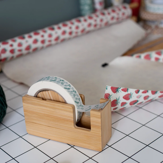 A roll of washi tape with a strawberry and blue floral pattern is displayed on a bamboo tape dispenser, with rolls of matching strawberry-patterned tape in the background on a white tiled surface.