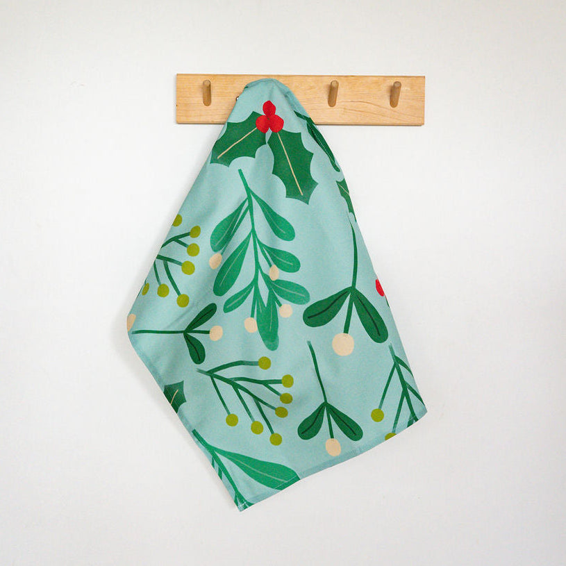 A green tea towel with a holly and mistletoe pattern hangs from a wooden peg rail on a white wall, with a blue-gray wall cabinet visible above.
