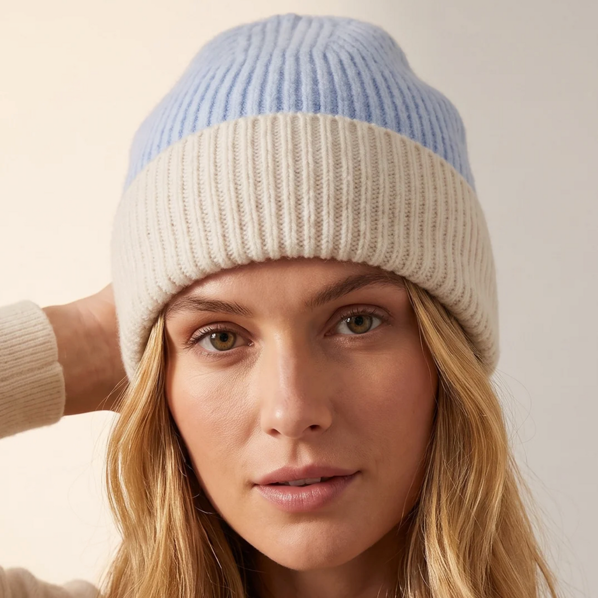 A close-up of a woman wearing a ribbed knit beanie with a two-tone design: light blue on top and cream on the lower half. The image is cropped from the chest up against a soft, light beige background.