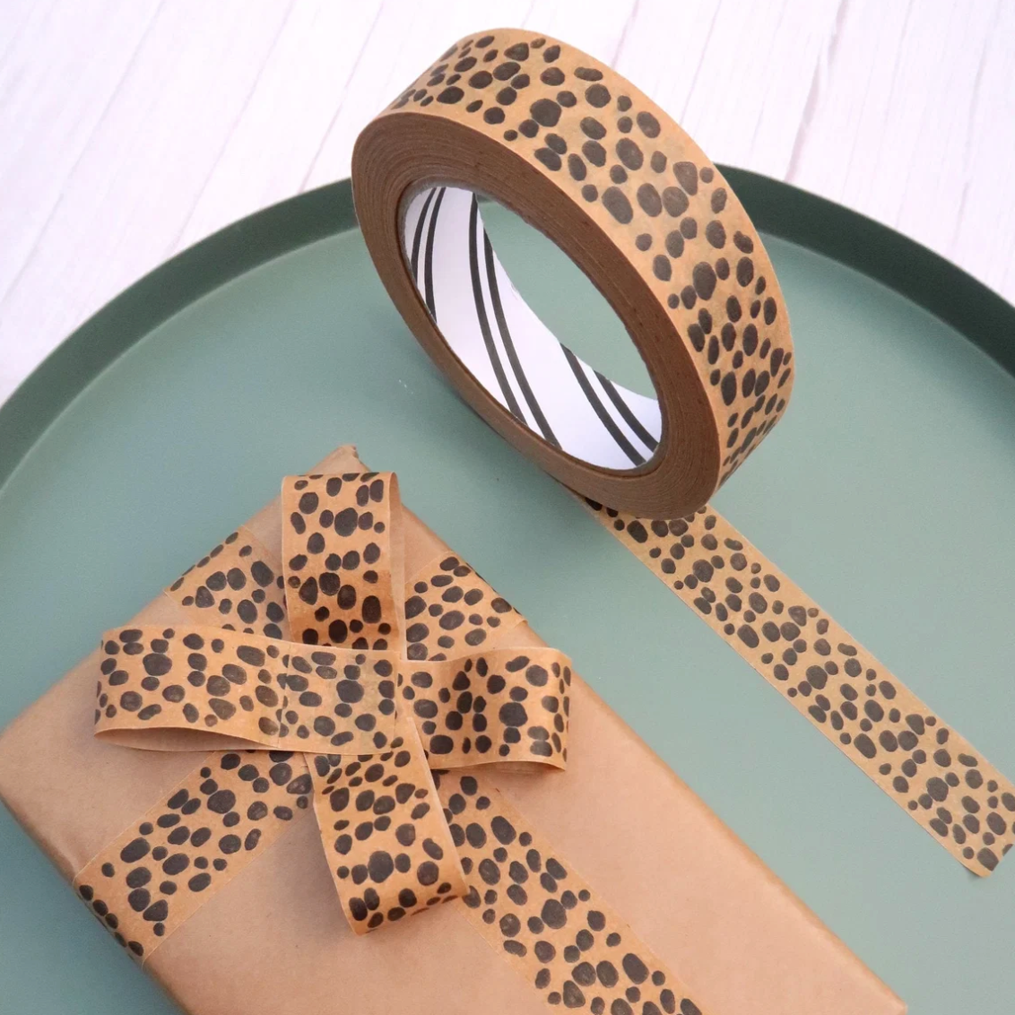 A roll of brown washi tape with a black leopard print pattern, shown partially unrolled next to a wrapped gift box tied with a matching ribbon on a round teal tray.