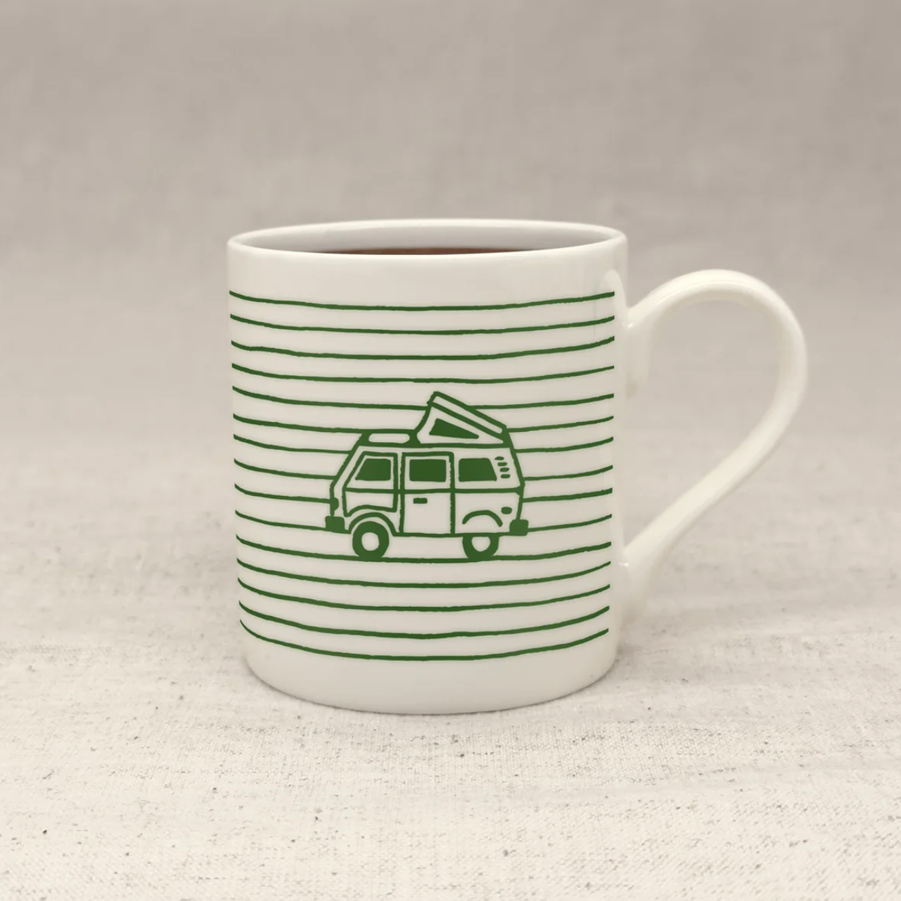 A white ceramic mug with thin green horizontal stripes and a green line-art illustration of a vintage camper van, shown on a light textured fabric surface against a neutral gray background.