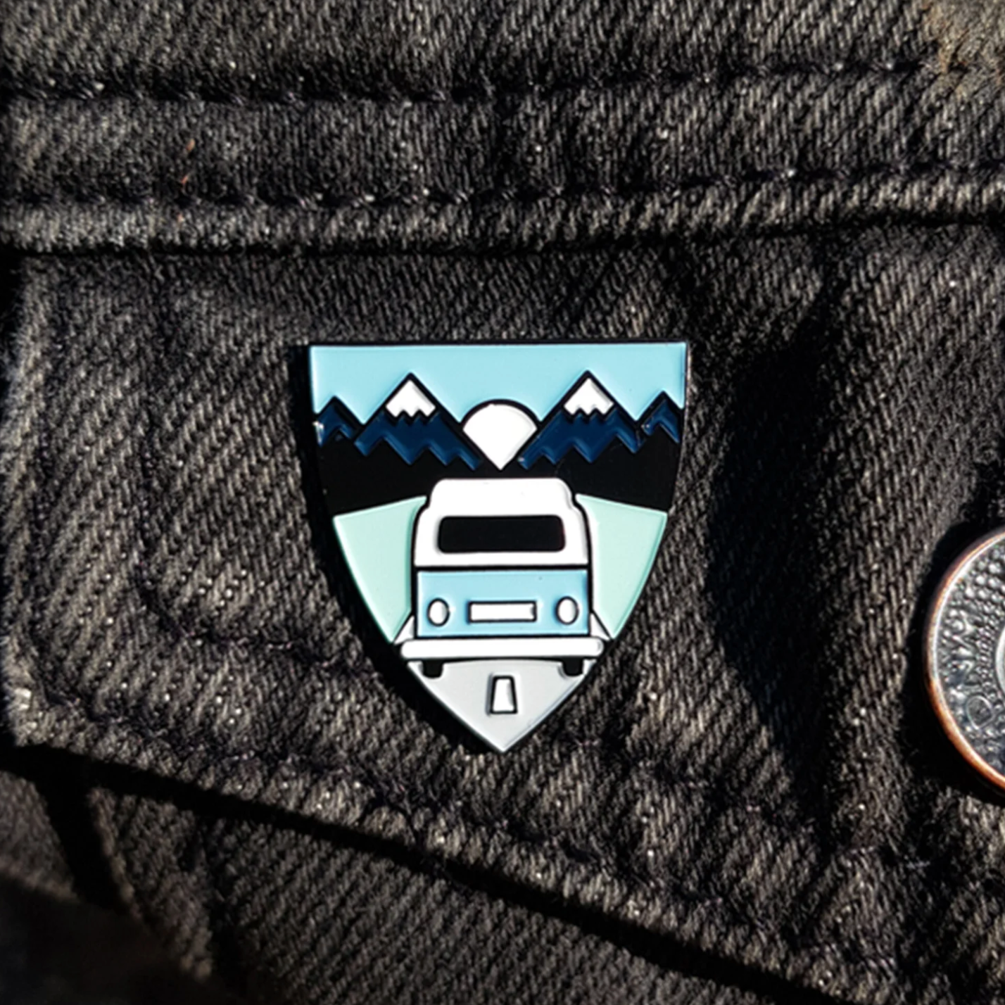 Pin with a design of a camper van and mountains on a dark fabric background