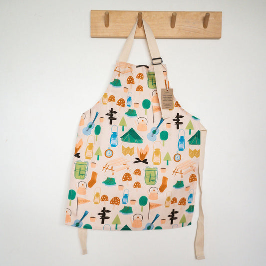 A cream-coloured children's kitchen apron with a colourful forest and camping pattern, hanging from a wooden peg on a white wall.