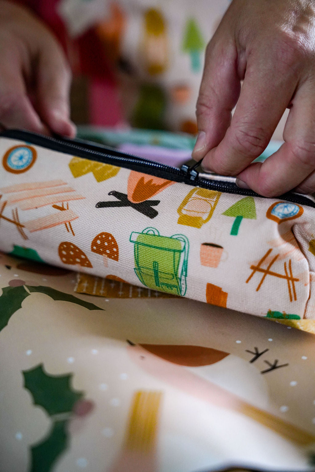 Close-up of a person's hands zipping a fabric pouch with a colorful camping-themed print on a cream background.