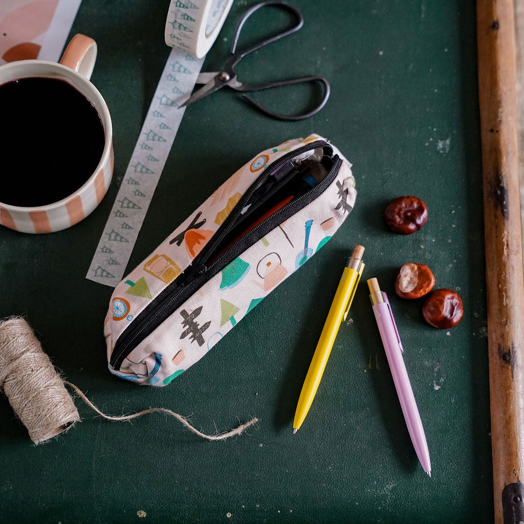 A patterned fabric pencil case with a black zipper lies on a dark green suitcase, surrounded by a coffee mug, scissors, measuring tape, pencils, and small decorative items.